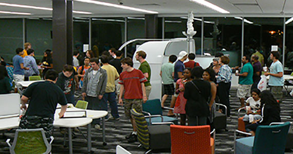 A group of young men and women stand around a large room with brightly colored chairs and tables